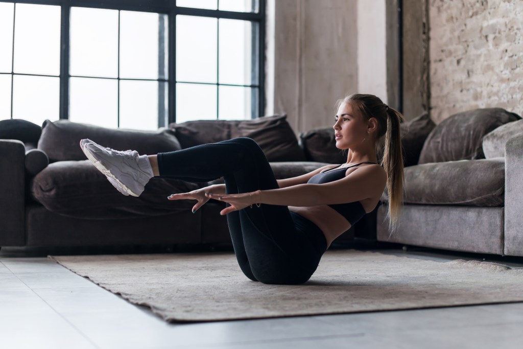 A woman performing a seated exercise on a rug in a modern living room, with legs raised and hands reaching forward.